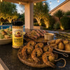 One bottle of BuffalowBBQ Cowboy Butter Seasoning on an outdoor kitchen counter beside grilled steak, shrimp skewers, asparagus, roasted potatoes and Caesar salad at dusk.