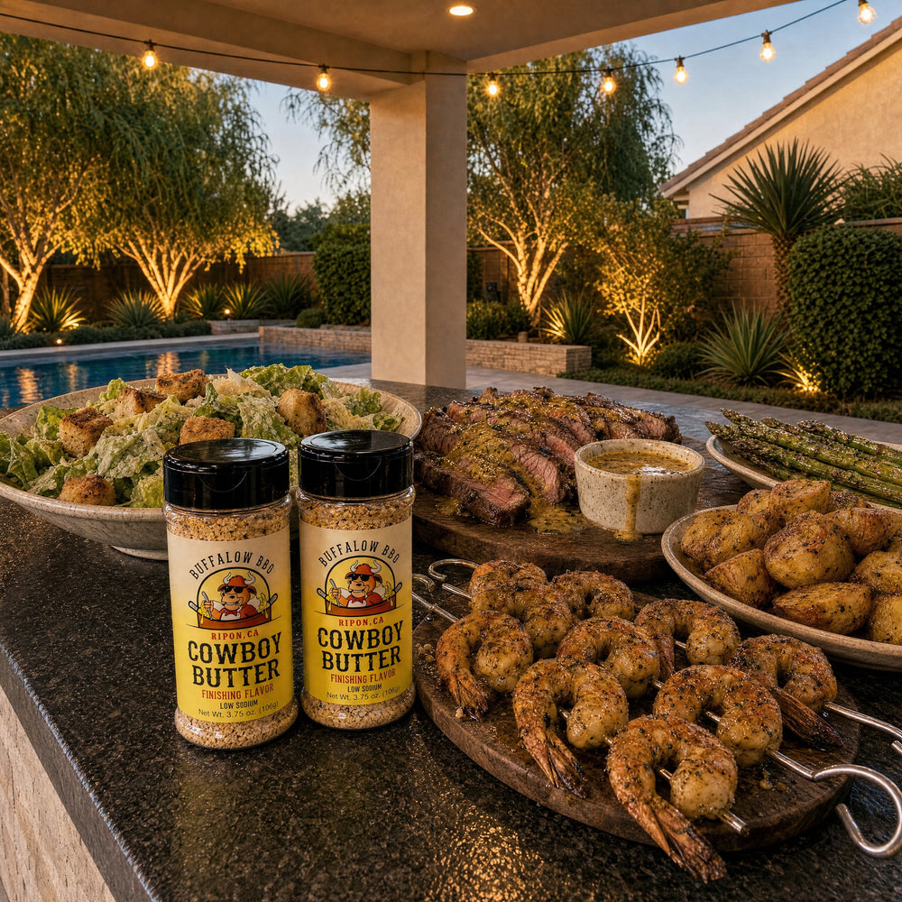 Two bottles of BuffalowBBQ Cowboy Butter Seasoning on an outdoor kitchen counter beside grilled steak, shrimp skewers, asparagus, roasted potatoes and Caesar salad at dusk.