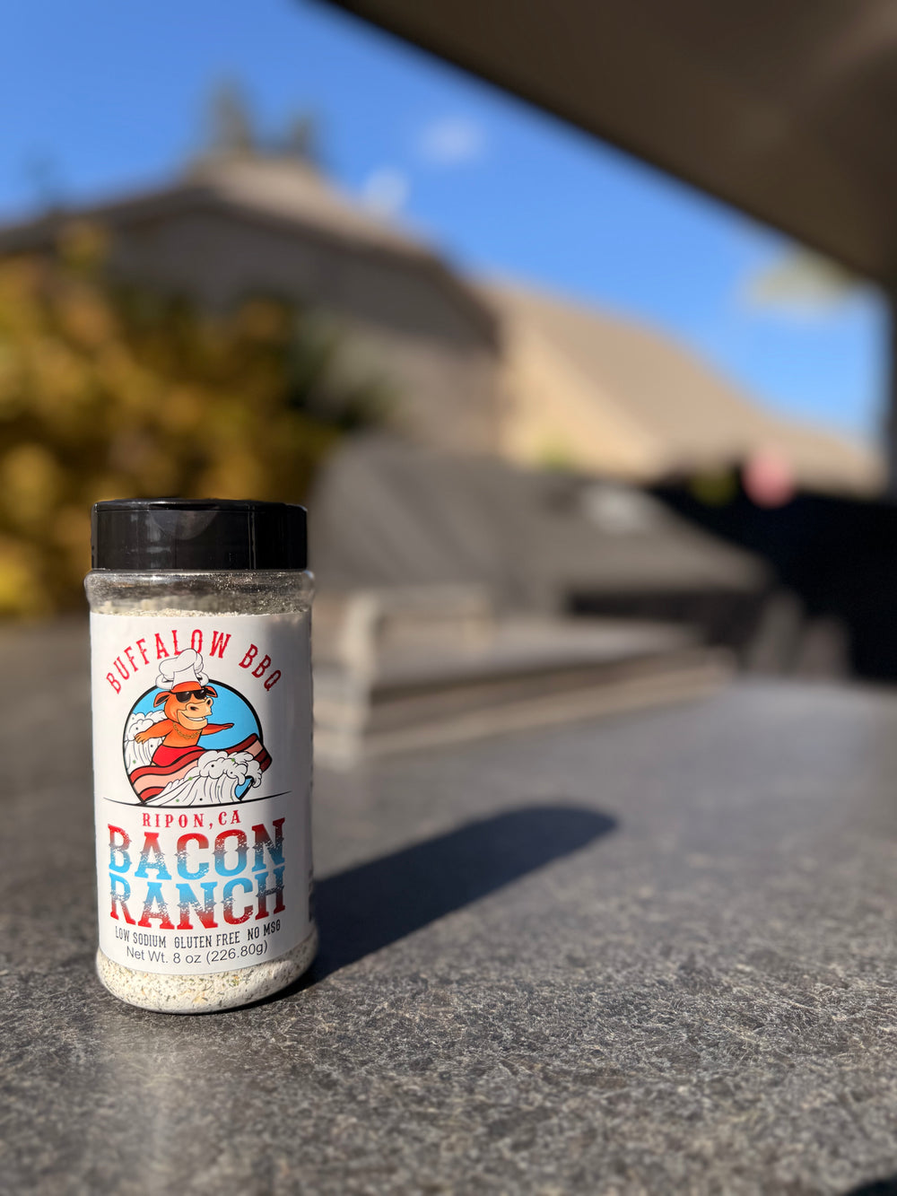 BuffalowBBQ Bacon Ranch all-purpose seasoning bottle on an outdoor countertop with a blurred backyard grilling scene.