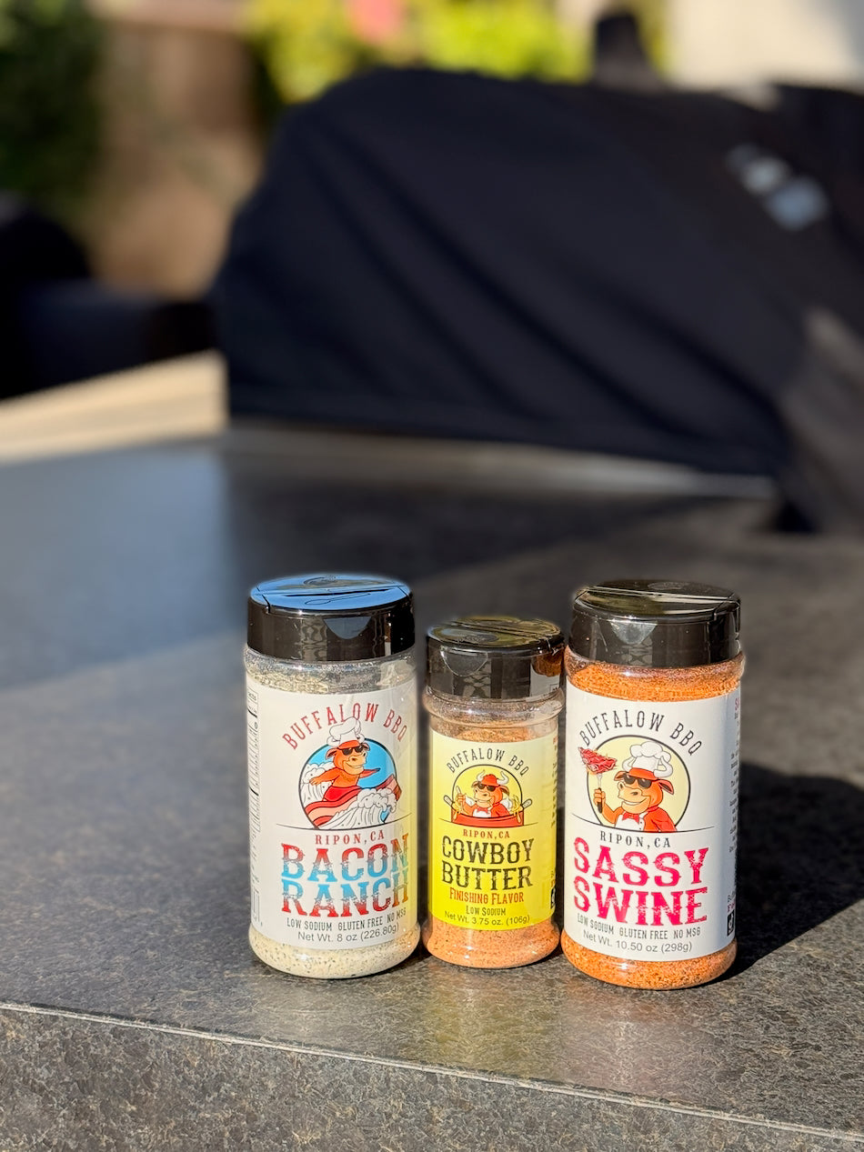 BuffalowBBQ Fan Favorites bundle featuring Bacon Ranch, Cowboy Butter, and Sassy Swine seasonings displayed together on an outdoor countertop with a blurred backyard grilling scene.